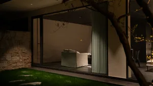 Exterior night view looking through large glass doors into a modern living room featuring a white sofa, textured stone wall, and warm ambient lighting.