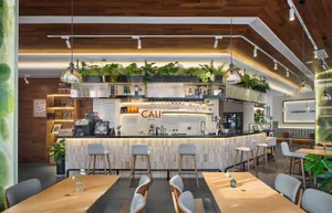 Modern cafe interior with a curved wooden bar counter, grey bar stools, abundant hanging plants, warm wood ceilings, and pendant lights.