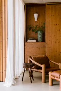 Modern minimalist living room corner featuring a brown leather and oak armchair, dark wood stump side table, built-in oak shelving with books and plants, white sheer curtain, and concrete ceiling.