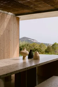 Speckled terrazzo counter with three rustic ceramic vases in beige, black and light terracotta, set against a wooden slat wall and panoramic mountain view under a thatched roof.