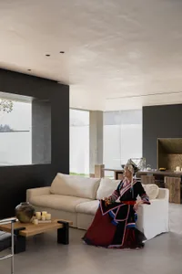 Woman wearing colorful traditional Taiwanese Indigenous dress and headdress sitting on a white sofa in a modern minimalist concrete living room with large windows.