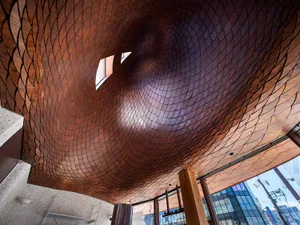 Looking up at a dramatic sculptural ceiling made of overlapping copper scales forming a swirling vortex pattern with small skylights visible.