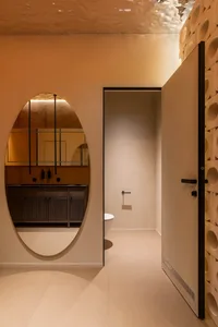 Modern minimalist bathroom with warm terracotta plaster walls, a large oval mirror reflecting a dark wood vanity, and a sculptural curved wall with circular cutouts.