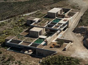 Aerial photograph of a modern stone courtyard villa with green infinity pools integrated into the arid Mediterranean hillside.