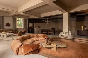 Spacious open-plan living room featuring a large curved terracotta velvet sofa, marble coffee tables, concrete pillars, and warm wood-paneled walls under a dramatic coffered ceiling.