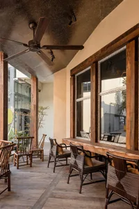 Covered outdoor patio featuring reclaimed wood beams, ceiling fan, large windows, and multiple dark rattan and wooden armchairs arranged along a long wooden counter.