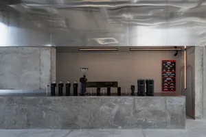 Modern minimalist coffee bar featuring a massive concrete counter, stainless steel backsplash with wavy texture, black coffee equipment, and a digital menu board under linear lighting.