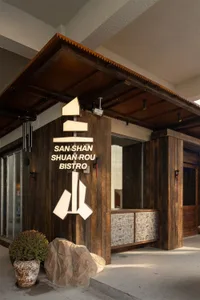 Exterior entrance of San Shan Shuan Rou Bistro featuring dark reclaimed wood cladding, illuminated three-mountain logo, stone planter, and textured stone base.