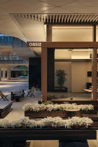 Modern courtyard lounge with corten steel planters filled with silvery white foliage, wooden benches, and a large glass-walled room in the background under a covered architectural ceiling.