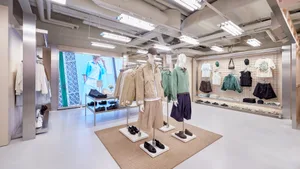 Modern minimalist clothing boutique interior with exposed concrete ceiling, industrial fluorescent lighting, steel display racks, and mannequins dressed in contemporary casual wear on a beige rug.