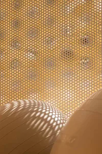 Close-up view of a light-colored wooden perforated screen with hundreds of uniform circular holes, with bright sunlight creating dotted shadow patterns on a curved wooden surface below.