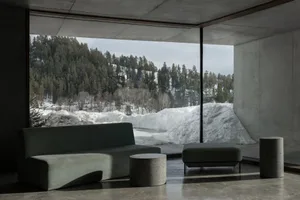 Modern minimalist living room with concrete walls and floor, featuring a curved green fabric sofa, matching ottoman, cylindrical concrete side tables, and a massive picture window overlooking a snowy forest and mountain.