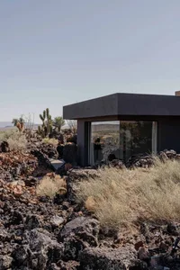 Modern concrete house with large glass windows situated among dark volcanic rocks and desert vegetation under a clear blue sky.