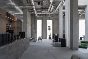Modern industrial cafe interior featuring exposed concrete walls and floors, large stainless steel ventilation ducts, a coffee counter with espresso machines, and minimalist furniture under bright natural light from tall windows.