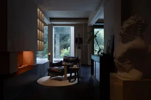 Modern minimalist living room featuring a built-in concrete fireplace with warm firelight, a black leather lounge chair on a round jute rug, a large marble torso sculpture in the foreground, and floor-to-ceiling windows overlooking a lush green forest.