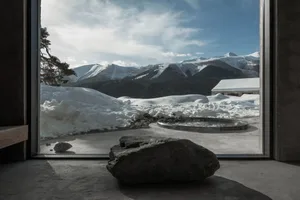 Interior view of a modern concrete room with a large glass window overlooking snow-covered mountains, a circular outdoor hot tub, and a large natural rock in the foreground.