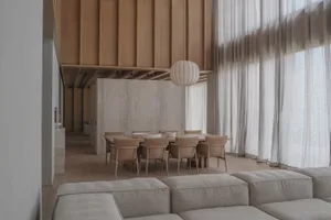 Modern minimalist dining area featuring a long oak table with beige upholstered chairs, a large paper lantern pendant light, concrete accent wall, and light oak flooring, viewed from a neutral-toned sofa.