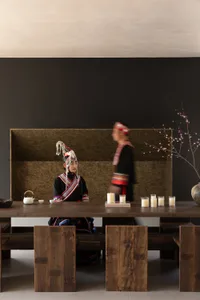 Woman in traditional Taiwanese indigenous clothing seated at a long reclaimed wood dining table with candles and tea set, another person in traditional attire walking in the background against a dark textured wall.