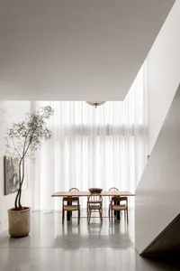 Minimalist dining room featuring a long oak wood table, four wooden chairs with woven seats, a large potted tree, sheer white curtains, and a woven pendant light.