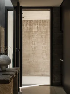 View from a minimalist entryway looking through an open black-framed door toward a large textured raw concrete wall bathed in natural light.