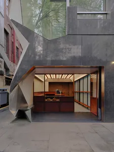 Modern bakery storefront featuring a dramatic angular facade made of weathered corten steel and concrete, with an open view into a wood-lined interior displaying fresh pastries.