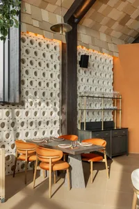 Modern restaurant dining area featuring white terracotta cinder block walls with integrated warm lighting, a dark wood dining table surrounded by four orange velvet chairs with wooden frames, a brass and black sideboard, and wooden plank ceiling.