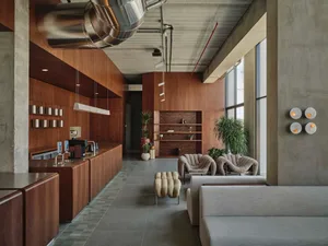 Modern industrial cafe lounge with extensive walnut wood cabinetry, concrete walls, large windows, boucle armchairs, and a prominent stainless steel ventilation duct suspended from the ceiling.