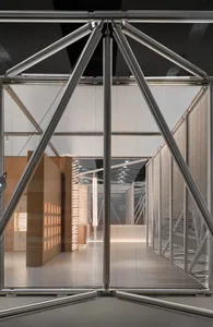 View through a large geometric stainless steel art installation with triangular trusses and horizontal blinds in the background.