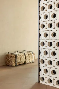 Modern minimalist entryway featuring a large off-white perforated terracotta screen, a black metal-framed bench with two cream sheepskin throws, and a bundle of dried reeds against a textured beige wall and large-format beige floor tiles.