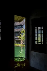 從昏暗室內透過石門望向鬱鬱蔥蔥的綠色水池庭院，遠處可見傳統中式建築與植物。