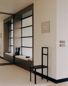 Modern minimalist entryway featuring a built-in dark wood window niche with white seat, a tall black metal chair with rivet details, and a framed abstract line drawing on a cream wall.
