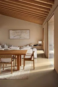 Modern minimalist dining area with solid oak table and woven rattan chairs on a textured rug, white sectional sofa in the background, exposed wooden beam ceiling and large abstract painting on the wall.