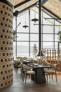 Modern restaurant interior with curved terracotta block column, dark wood dining tables, cream upholstered chairs, large windows showing snowy mountain view, and wooden ceiling.