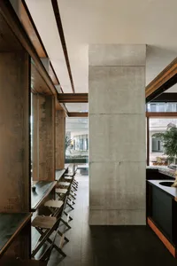 Narrow industrial cafe interior featuring a massive raw concrete pillar, long wooden counter with folding wooden stools, dark metal accents and natural light from large windows.
