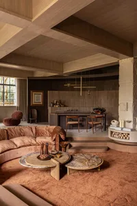 Spacious living room featuring a curved terracotta velvet sofa, two round marble coffee tables, wood-paneled walls and ceiling, and a raised dining area with wooden chairs in the background.