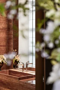 Close-up of a luxurious brown marble bathroom vanity with integrated rectangular sinks and two modern chrome faucets, reflected in a large mirror with white floral arrangements in the foreground.