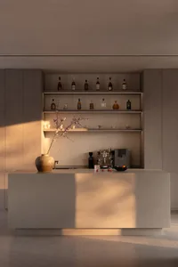 Modern minimalist concrete kitchen island used as a home bar with backlit floating shelves displaying liquor bottles and tableware, featuring a large ceramic vase with flowering branches in soft natural light.