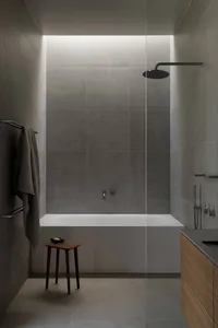 Modern minimalist bathroom featuring large-format concrete tiles, a white freestanding bathtub, black rain shower, wooden vanity, and a small wooden stool with a towel hanging on a black rack.
