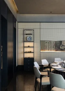 Modern dining room featuring a backlit perforated panel wall, built-in display shelving with blue and white porcelain, a large Chinese calligraphy artwork, and white upholstered dining chairs around a dark table.