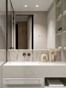 Modern minimalist bathroom vanity with white concrete countertop, integrated sink, marble backsplash, chrome wall-mounted faucet, and decorative travertine sculptures on open shelving.