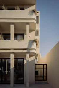 Exterior view of a modern multi-story concrete building featuring rounded balconies, vertical pillars, large glass doors, and potted grasses under a clear blue sky.