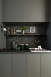 Modern sage green kitchen cabinetry with dark green marble backsplash and countertop, open shelf displaying books, plants, and decor, white ceramic sink with matte faucet.