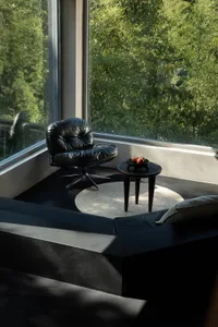 Modern black leather lounge chair with chrome base placed on a circular jute rug next to a dark wood side table holding red apples, set in a sunlit corner with floor-to-ceiling windows overlooking dense green bamboo forest.