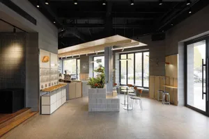 Modern industrial-style cafe interior with grey concrete tiles, light wood accents, central planter, and round seating area by large windows.