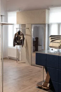 Modern minimalist entryway featuring a tall fluted stone wardrobe with integrated mirror, a blue upholstered console table, and a leather jacket hanging on a chrome rack against a light wood floor.