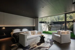 Modern bedroom featuring a platform bed with dark headboard, two white sofas, a patterned area rug, and large floor-to-ceiling windows overlooking a landscaped garden with stone walls.