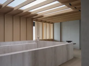 Modern minimalist living room featuring massive travertine blocks forming sunken seating, light wood ceiling beams, and long rectangular skylights allowing natural daylight.