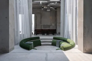 Modern brutalist interior featuring a large curved emerald green velvet sofa arranged in a sunken circular platform, surrounded by sheer white curtains and exposed concrete walls with visible ductwork.