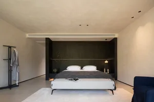 Modern minimalist bedroom with dark wood panel headboard wall, beige textured plaster walls, white bedding, upholstered bench, and blue velvet armchair