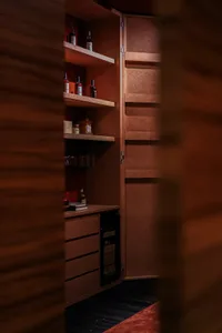 Interior view of a luxurious home bar cabinet made of burl wood with open shelves holding liquor bottles, glassware, a mini fridge, and warm LED lighting.
